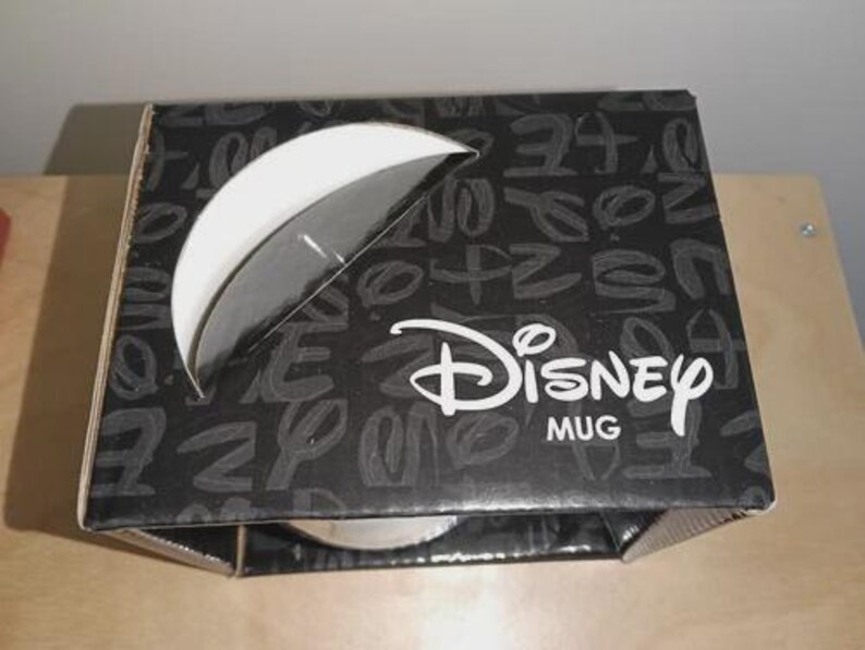 May include: A black and white Disney mug box with a partial view of the mug inside. The box has a white semi-circle cutout and the Disney logo in white. The box is covered in a pattern of white letters.