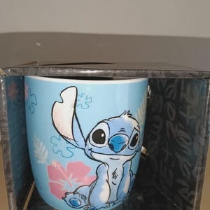 May include: A blue ceramic mug featuring the character Stitch from Lilo & Stitch. The mug has a light blue background with pink hibiscus flowers. Stitch is depicted in a seated position with a smiling expression. The mug is in a black box.