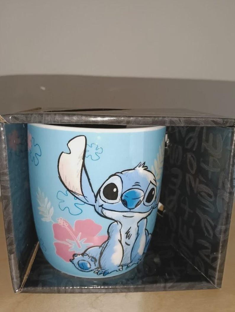May include: A blue ceramic mug featuring the character Stitch from Lilo & Stitch. The mug has a light blue background with pink hibiscus flowers. Stitch is depicted in a seated position with a smiling expression. The mug is in a black box.