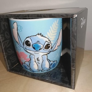 May include: A light blue ceramic mug featuring the character Stitch from Disney's Lilo & Stitch. The mug is decorated with a smiling Stitch, pink flowers, and green leaves. The mug is packaged in a black box with the Disney logo.