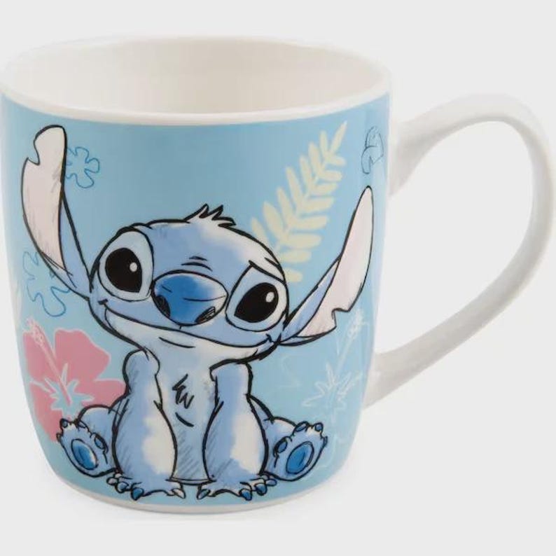 May include: A light blue ceramic mug featuring a cartoon character, Stitch, with large black eyes and blue accents. The mug has a white handle and interior, with floral and leaf designs. The mug is a standard size for hot beverages.