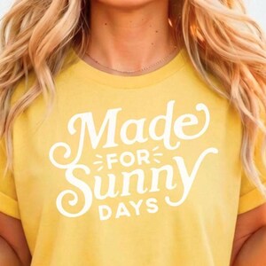 May include: A mustard yellow short-sleeved t-shirt featuring white screen-printed text on the front that reads Made FOR Sunny DAYS in a combination of cursive and block lettering. The shirt has a crew neckline and rolled sleeve cuffs.