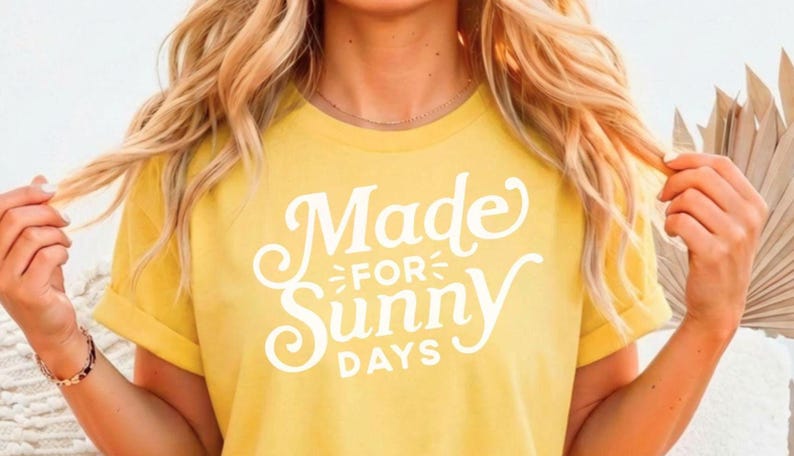 May include: A mustard yellow short-sleeved t-shirt featuring white screen-printed text on the front that reads Made FOR Sunny DAYS in a combination of cursive and block lettering. The shirt has a crew neckline and rolled sleeve cuffs.