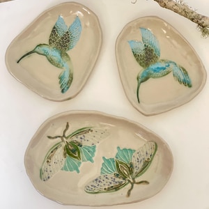 Puede incluir: Tres platos de cerámica con diseños de colibríes y polillas. Dos platos presentan colibríes con alas azules y verdes, mientras que el tercero muestra dos polillas con paletas de colores similares. Los platos tienen un fondo beige.