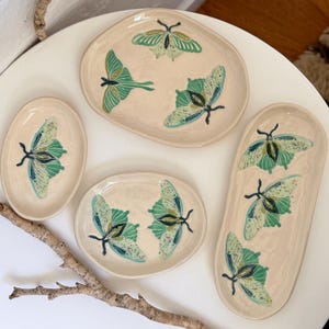 May include: A set of five ceramic serving dishes in different shapes. Each dish is off-white and features a green and blue butterfly design. The dishes are arranged on a white surface, with a wooden branch in the foreground.