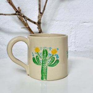 May include: A cream-colored ceramic mug featuring a hand-painted green cactus design with yellow flowers and small blue accents. The mug has a smooth, glossy finish and a curved handle on the left side, showcasing a detailed botanical illustration.