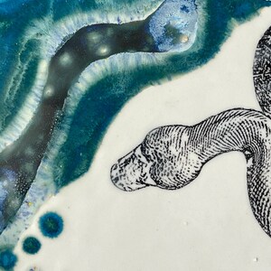 May include: A ceramic plate with a white surface, decorated with a black and white snake illustration. The upper left corner has an abstract design in shades of blue and teal, with a glossy finish.