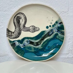 May include: A ceramic plate featuring a black and white snake illustration. The plate has a white background with a turquoise and blue wave design on the lower half. A decorative piece for the home.