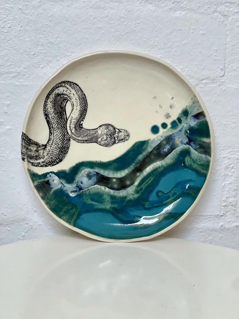 May include: A ceramic plate featuring a black and white snake illustration. The plate has a white background with a turquoise and blue wave design on the lower half. A decorative piece for the home.