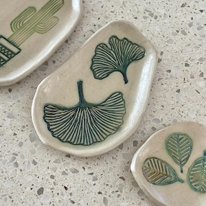 Könnte beinhalten: Eine Sammlung kleiner, cremefarbener Keramikschalen mit botanischen Designs. Eine Schale zeigt einen grünen Kaktus, eine andere zwei Ginkgoblätter. Die dritte Schale zeigt drei Blätter. Die Designs sind geätzt und mit grüner Glasur gefüllt.