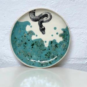 May include: A ceramic plate with a white centre and a turquoise blue rim. A black and white illustration of a snake is in the centre. The plate has a glossy finish and is likely handmade.
