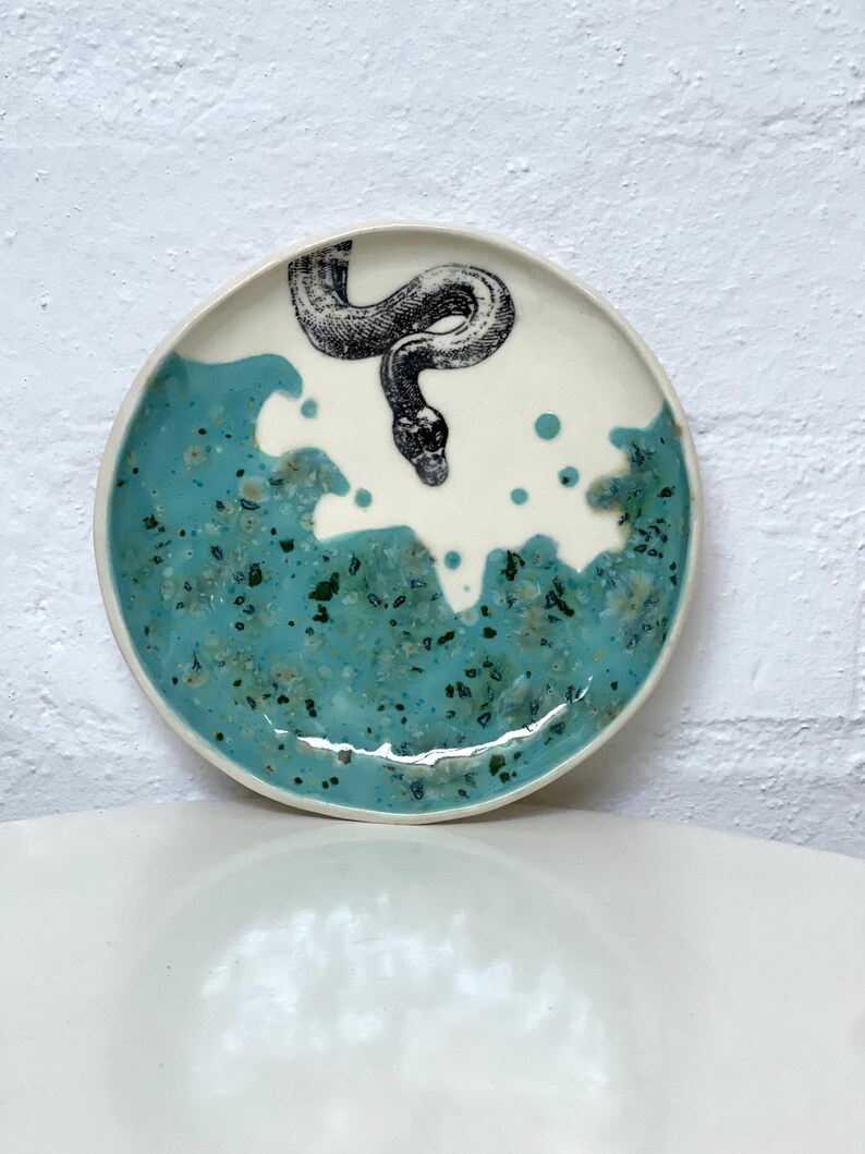 May include: A ceramic plate with a white centre and a turquoise blue rim. A black and white illustration of a snake is in the centre. The plate has a glossy finish and is likely handmade.