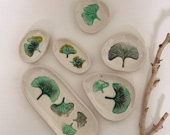 Puede incluir: Un conjunto de seis platos de cerámica en color blanco roto con formas irregulares, cada uno con diseños de hojas de ginkgo pintados a mano en verde. El set incluye una bandeja ovalada larga, cuatro platos pequeños redondeados y uno más grande.