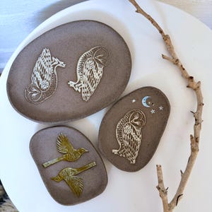 May include: Three oval-shaped ceramic dishes with a matte brown finish. The largest dish features two white owl illustrations. A smaller dish displays one white owl with a crescent moon and stars. The third dish shows two golden-yellow birds in flight.