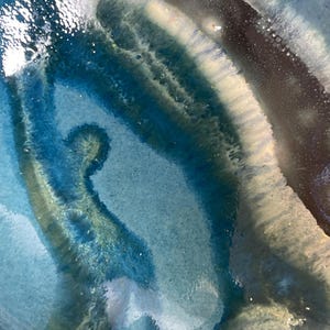 May include: Close-up of a ceramic piece with a glossy, layered design. The artwork features shades of blue, teal, and white, creating a fluid, abstract pattern. The edges of the piece are a creamy off-white colour.