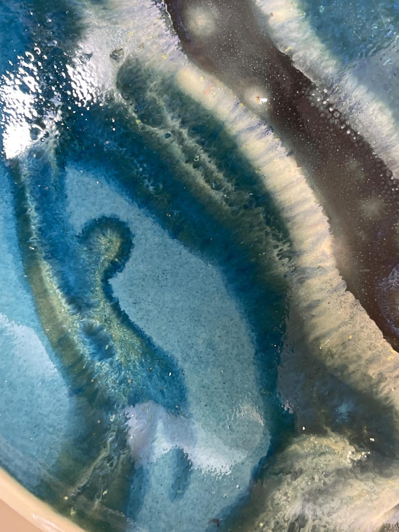 May include: Close-up of a ceramic piece with a glossy, layered design. The artwork features shades of blue, teal, and white, creating a fluid, abstract pattern. The edges of the piece are a creamy off-white colour.