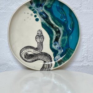 May include: A ceramic plate with a cream base and a turquoise and blue glazed rim. A detailed black and white illustration of a snake is on the plate. The plate has a glossy finish and is likely handmade.