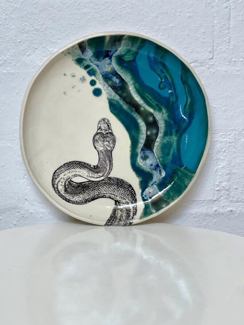 May include: A ceramic plate with a cream base and a turquoise and blue glazed rim. A detailed black and white illustration of a snake is on the plate. The plate has a glossy finish and is likely handmade.