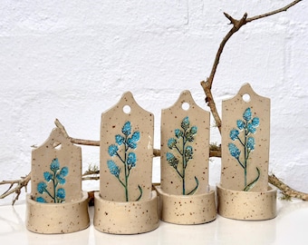 Può includere: Quattro segnaposto per piante in ceramica con finitura beige macchiettata. Ogni segnaposto presenta un motivo floreale blu e un piccolo foro nella parte superiore. I segnaposto sono inseriti in basi beige abbinate, con un rametto naturale sullo sfondo.