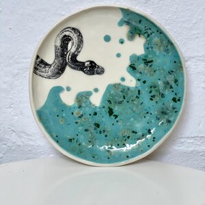 May include: A round ceramic plate with a white and turquoise design. A black and white illustration of a snake is on the white portion of the plate. The turquoise section has a speckled pattern.
