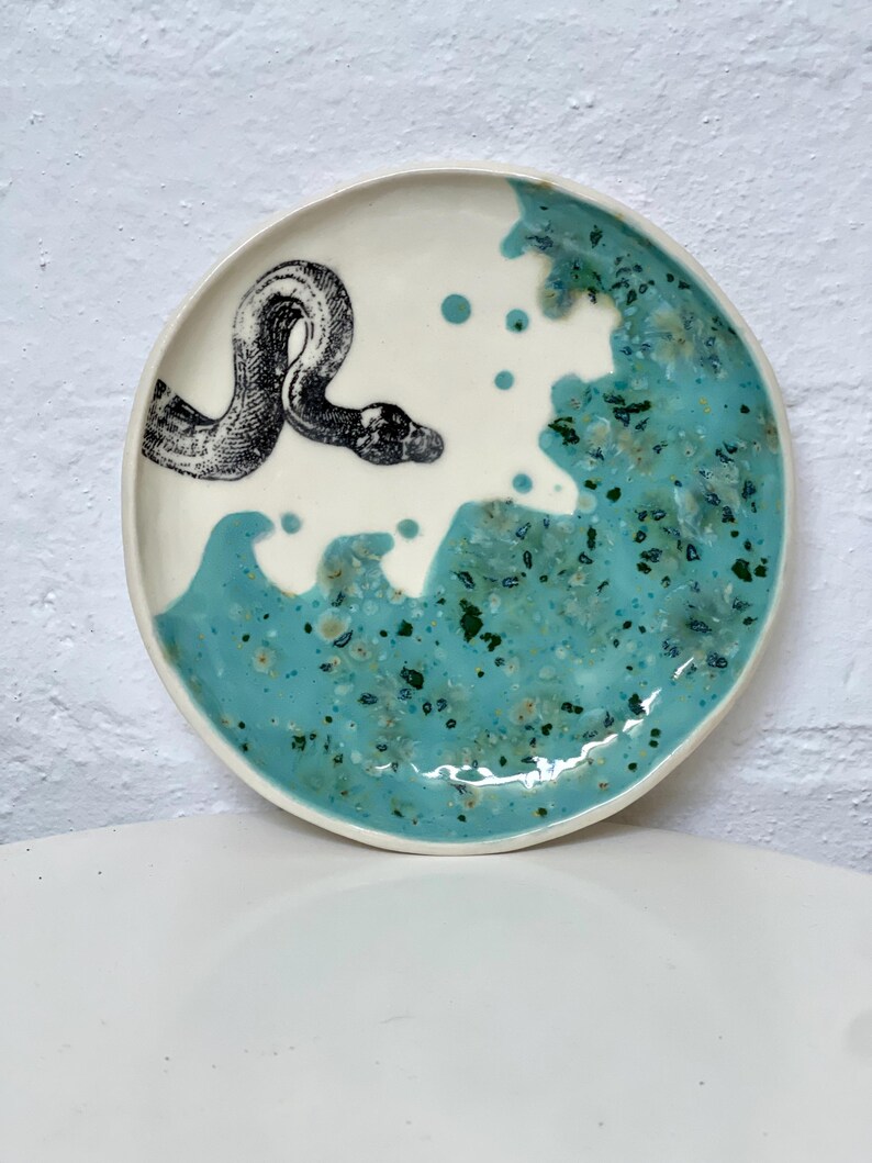 May include: A round ceramic plate with a white and turquoise design. A black and white illustration of a snake is on the white portion of the plate. The turquoise section has a speckled pattern.