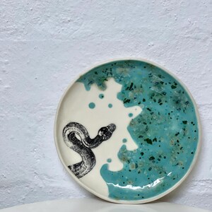 May include: A circular ceramic plate featuring a white base and a turquoise blue glaze. A black and white illustration of a snake is on the white section. The plate has a slightly raised edge.