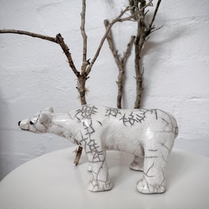 Könnte beinhalten: Eine weiße Eisbär-Figur aus Keramik mit schwarzen Craquelé-Details. Der Bär steht auf einer weißen Oberfläche, im Hintergrund kahle Äste. Die Figur hat eine glänzende Oberfläche und ist ein dekorativer Artikel.