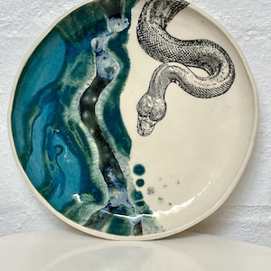 May include: A ceramic plate with a cream background and a teal and blue abstract design on the left. A black and white illustration of a snake is on the right side of the plate.