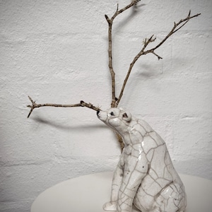 Könnte beinhalten: Eine weiße Keramik-Eisbärfigur mit Rissglasur-Effekt, sitzend und nach oben blickend. Ein kleiner, kahler Ast erstreckt sich von hinter dem Bären. Die Figur steht auf einer weißen Oberfläche vor einer weißen Backsteinmauer.