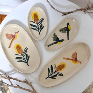 May include: Four oval-shaped ceramic dishes in a cream colour, each featuring hand-painted illustrations. Two dishes display a yellow and orange banksia flower, while two feature birds in flight with yellow and brown plumage and dark green wings.