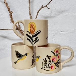 May include: Three cream-coloured ceramic mugs stacked together. The top mug features a yellow and orange banksia flower. The bottom left mug shows a yellow and black bird in flight. The bottom right mug displays pink and yellow flowers with green leaves.