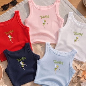 May include: Five ribbed tank tops in red, light pink, white, navy blue, and light blue. Each sleeveless shirt features a graphic of the character Tinker Bell and the text TinkerBell printed in green on the center of the chest area.