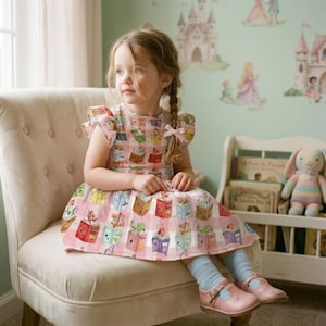 Puede incluir: Una niña pequeña sentada en una silla color crema, lleva un vestido rosa con un estampado de libros. El vestido presenta un diseño a cuadros con ilustraciones de libros abiertos. Lleva zapatos rosas y calcetines azul claro. Un conejito de peluche y libros en el fondo.