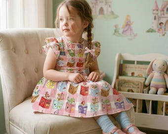 May include: A young girl sits in a cream-colored chair, wearing a pink dress with a book-themed pattern. The dress features a checkered design with illustrations of open books. She wears pink shoes and light blue socks. A stuffed bunny and books are in the background.