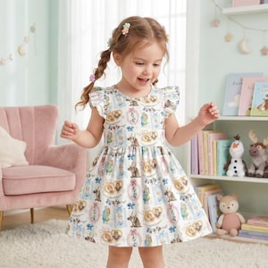 Può includere: Una bambina indossa un vestito bianco con un motivo colorato con personaggi del film "Frozen". L'abito ha maniche corte con volant e una gonna svasata. Lo sfondo include una sedia rosa e una libreria con libri e giocattoli.