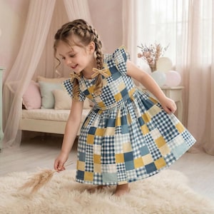 Op de afbeelding: Een jong meisje draagt een blauwe, gele en witte geruite jurk met korte mouwen en een uitlopende rok. De jurk heeft een patchwork-ontwerp. Het meisje is blootsvoets en houdt een gedroogde plant vast. De achtergrond omvat een bed, gordijnen en ballonnen.