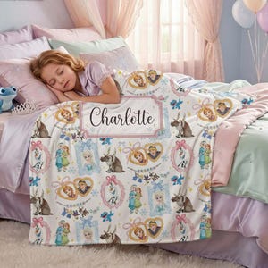 Könnte beinhalten: Eine weiße Fleece-Decke mit Figuren aus dem Film Frozen, darunter Anna, Elsa, Olaf und Sven. Die Decke hat einen rosa Rand und einen personalisierten Namen, "Charlotte", in einem dekorativen Rahmen. Die Decke liegt auf einem Bett mit rosa und lila Bettwäsche.