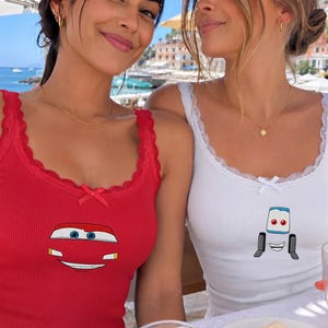 May include: A red ribbed tank top featuring a graphic of a car front with blue eyes and a white smile, and a white ribbed tank top featuring a graphic of a forklift character with blue eyes and a wide smile, both with lace trim and a small bow.