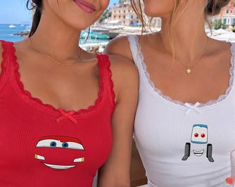 May include: A red ribbed tank top featuring a graphic of a car front with blue eyes and a white smile, and a white ribbed tank top featuring a graphic of a forklift character with blue eyes and a wide smile, both with lace trim and a small bow.