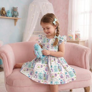 May include: A young girl wearing a white dress with a colorful monster print, holding a blue plush toy. The dress has flutter sleeves and a round neckline. The girl is sitting on a pink sofa in a room with monster-themed decor.
