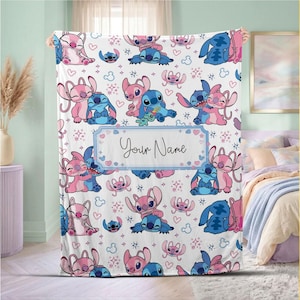 May include: A white blanket featuring a repeating pattern of blue and pink cartoon characters. The blanket has a rectangular label with the text "Your Name" in a script font. Hearts and stars are scattered across the design.