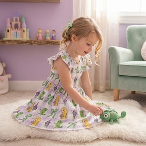 May include: A young girl wearing a light purple dress with a pattern of cartoon characters, bows, and castles. The dress has short sleeves and a flared skirt. She is playing with a green crocheted chameleon toy on a white rug.