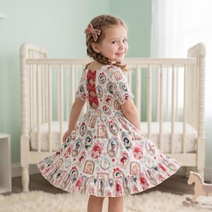 May include: A white dress with a princess-themed print, featuring a ruffled hem and short sleeves. The dress has a unique open back design with red bows. The girl is wearing a pink bow in her hair.