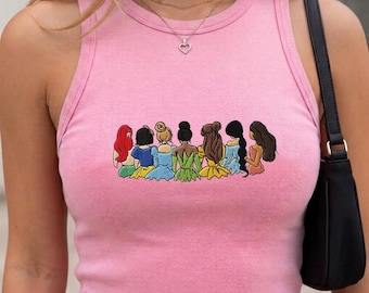 May include: A light pink sleeveless tank top featuring a graphic print of seven cartoon characters standing in a row, each with distinct hair colors and dresses. The characters are shown from the back, displaying various hairstyles and colorful outfits.