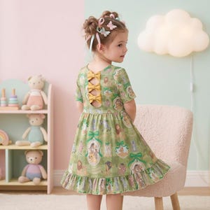 May include: A green dress with a repeating pattern of framed illustrations and a ruffled hem. The dress features a cut-out back with three yellow bows. The girl's hair is styled with ribbons and butterfly clips.