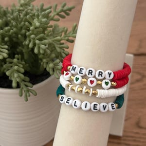 May include: Stack of three beaded bracelets with white letter beads spelling "MERRY" and "BELIEVE". The bracelets are red, white, and green with gold accents. A small green plant in a white pot is in the background.
