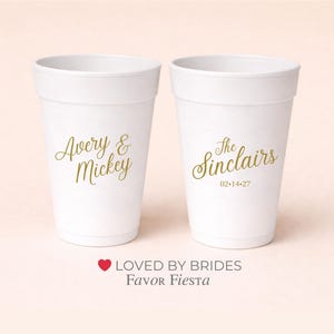 May include: Two white disposable cups with gold lettering. One cup reads "Avery & Mickey", the other reads "The Sinclairs 02-14-27". The cups are likely for a wedding or party.
