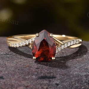 Pode incluir: Um anel de ouro com uma pedra preciosa vermelha escura em forma de pêra. A banda é adornada com pequenos diamantes brilhantes, colocado sobre uma superfície escura e texturizada.