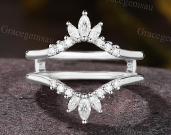 May include: A silver ring guard with a crown design. The ring features marquise-cut and round-cut clear gemstones. The ring guard is designed to sit alongside an engagement ring, enhancing its appearance.