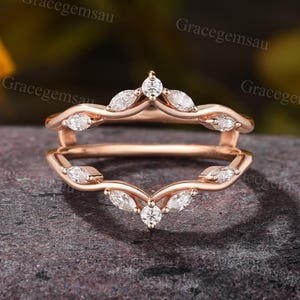 Op de afbeelding: Een roségouden ringbeschermer met een gebogen ontwerp. De ring heeft meerdere heldere, marquise-geslepen en rondgeslepen edelstenen. De ring is ontworpen om om een andere ring te zitten.